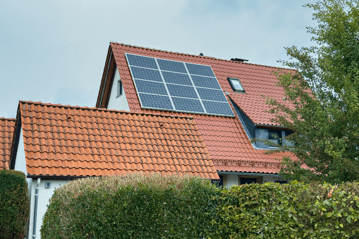 Residential house with red tile roof fitted with rooftop solar panels, partially framed by green hedges and trees under a cloudy sky.