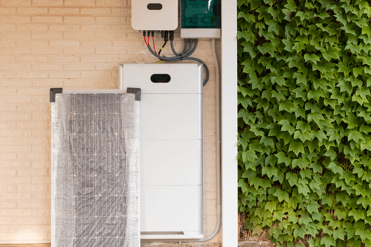 Residential solar battery storage system and inverter mounted on an exterior brick wall, with wiring visible and green ivy growing along the side.