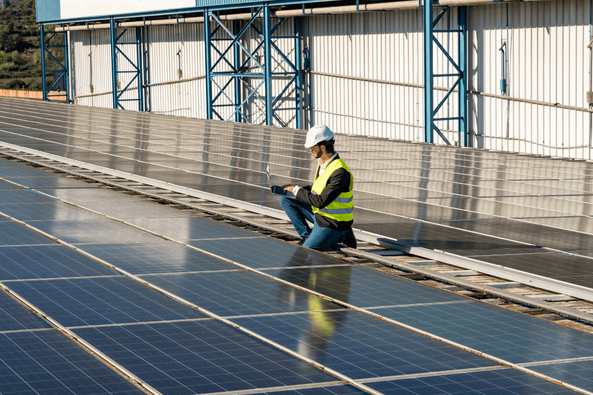 Technician wearing a hard hat and safety vest kneels on a commercial rooftop solar array, using a laptop to inspect rows of solar panels installed on an industrial building.